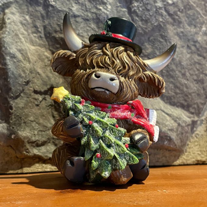 A Highland cow figurine wearing a black top hat and a red bow tie, holding a decorated green Christmas tree, set against a rustic stone wall background available at Rustic Ranch furniture in Airdrie