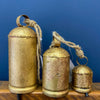 Three gold-colored bell-shaped ornaments with ropes against a blue background. Gold Clapper Bells - Three Sizes available at Quilted Cabin Home Decor.