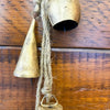 Two rustic gold bells hanging on a wooden surface. Mixed Gold Bells Hanger available at Quilted Cabin Home Decor.