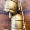 Two rustic gold bells hanging on a wooden surface with twine. Three Gold Clapper Bell Hanger available at Quilted Cabin Home Decor.