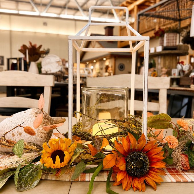 A rustic white metal lantern centerpiece with a clear glass jar candle holder, styled with orange sunflowers, dried leaves, and a plaid runner in a warm indoor setting.  Available at Quilted Cabin in Airdrie