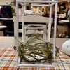 A rustic white wooden lantern with a clear glass jar and dried green mossy branches, displayed on a plaid table runner in a shop setting. Candle Ring available at Quilted Cabin in Airdrie