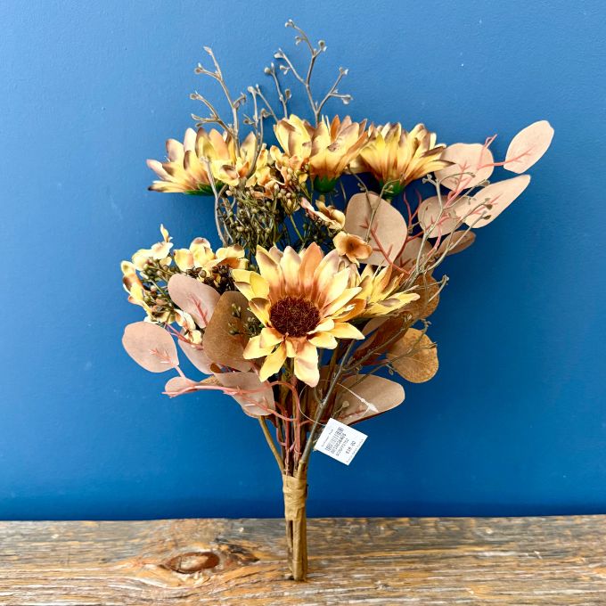A dried floral arrangement featuring yellow sunflowers, beige eucalyptus leaves, and brown branches, bundled in a brown paper sleeve and standing on a rustic wooden surface against a solid blue background.  Sunflower florals available at Quilted Cabin in Airdrie