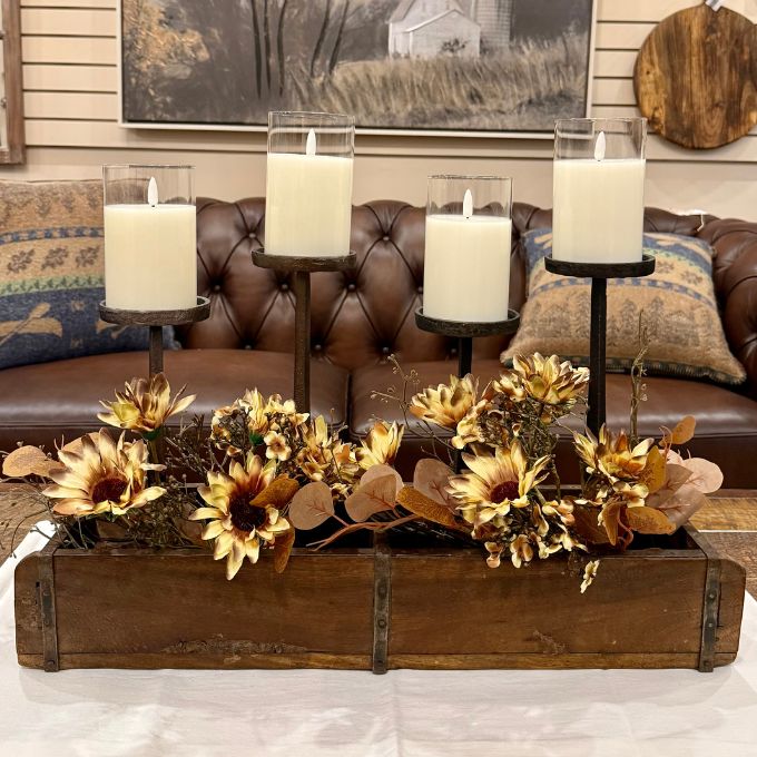 A rustic fall centerpiece arrangement in a wooden rectangular box, featuring dried sunflowers, eucalyptus leaves, and three black metal candle holders with white pillar candles, set in front of a brown leather tufted sofa.  All items available at Rustic Ranch Furniture and Quilted Cabin in Airdrie