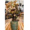 An artificial autumn floral arrangement in a speckled teal ceramic pot, featuring dark brown pine needles, yellow berries, acorns, and green leaves, displayed on a woven placemat in a dining room setting.