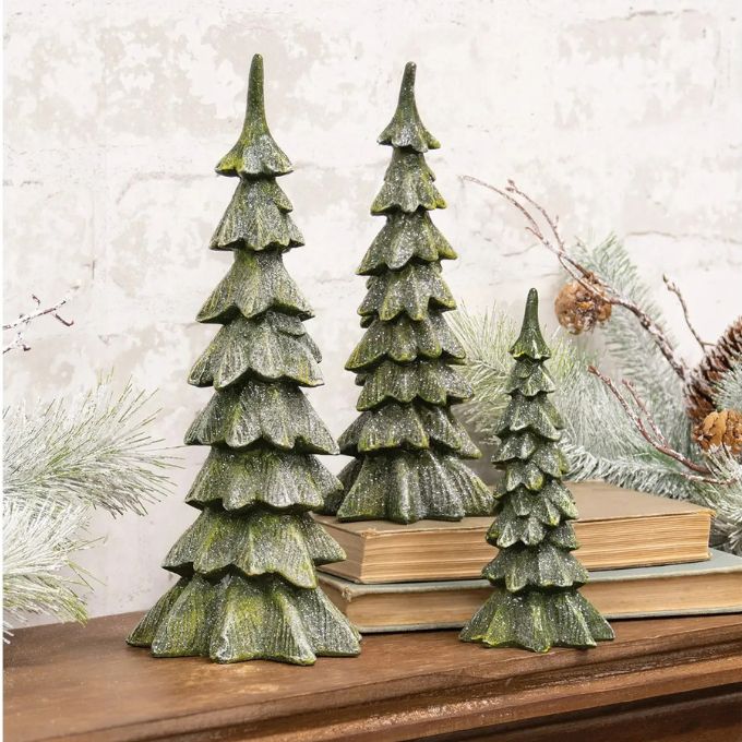 Three tiered pine tree ornaments in a textured dark green finish, displayed on a wooden mantel with books and holiday greenery.  Small and large trees available at Quilted Cabin in Airdrie