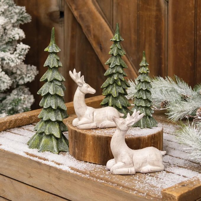 Two small off-white deer figurines posed among three green pine tree ornaments on a rustic wooden surface with faux snow and holiday greenery.  Small and large trees available at Quilted Cabin in Airdrie