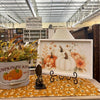 A framed autumn watercolor art print featuring white pumpkins, orange pumpkins, and orange flowers, displayed on a black stand on a floral table runner inside a store. All items available at Quilted Cabin in Airdrie