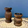 Two dark brown wooden candle holders with a textured, distressed finish, displayed on a patterned surface against a solid blue background.  Candle holders available at Quilted Cabin in Airdrie