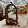 A rustic fall-themed centerpiece featuring a dark wood arched mirror with two white pillar candles inside, decorated with faux autumn leaves. It is styled on a red and white plaid runner next to a white textured ceramic vase filled with dried flowers, set on a white table inside a home decor store.  All Decor items available at Quilted Cabin Home Parties