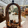 A rustic wooden arched decorative lantern centerpiece on a table in a home decor store. The lantern contains two white pillar candles and is surrounded by faux autumn leaves in orange, brown, and beige tones. All Decor items are available at Quilted Cabin in Airdrie