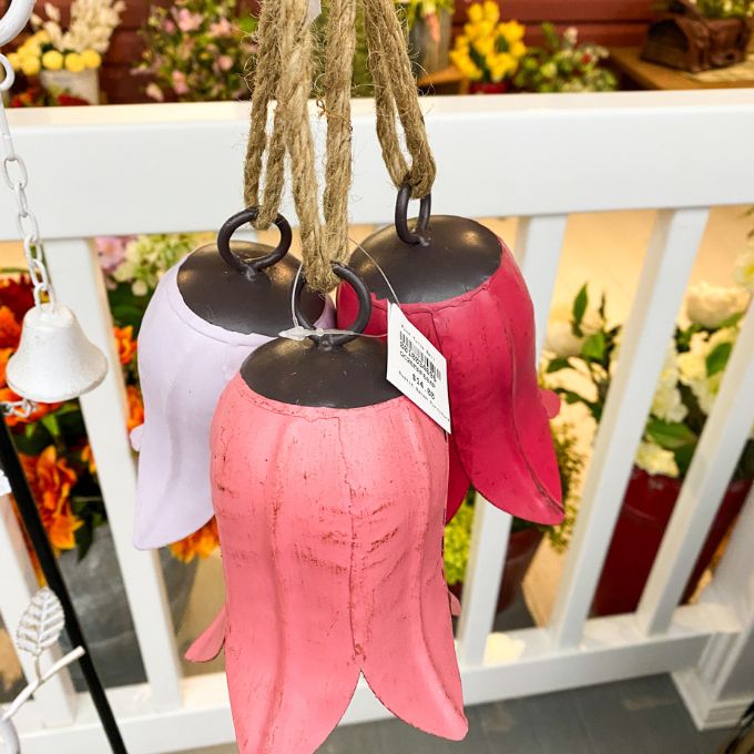 Pink and white wind chimes hanging on a white railing with a blurred floral background. Tulip Bells - Three Colours available at Quilted Cabin Home Decor.