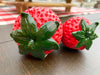 Two red strawberry-shaped objects with green leaves on a wooden surface. Resin Strawberries - Set of Two available at Quilted Cabin Home Decor.