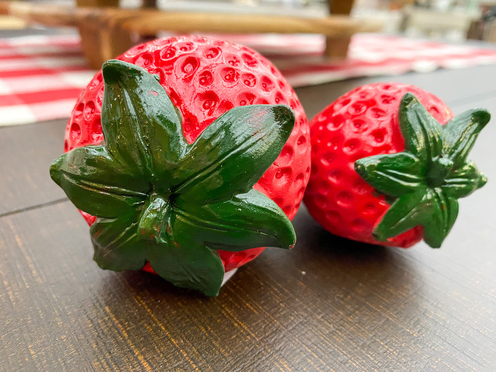 Two red strawberry-shaped objects with green leaves on a wooden surface. Resin Strawberries - Set of Two available at Quilted Cabin Home Decor.
