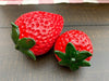 Two red strawberry-shaped objects with green leaves on a wooden surface. Resin Strawberries - Set of Two available at Quilted Cabin Home Decor.