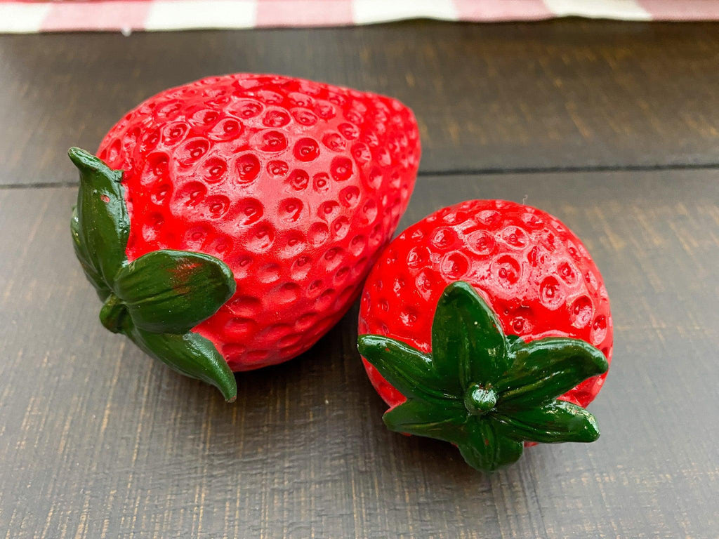 Two red strawberry-shaped objects with green leaves on a wooden surface. Resin Strawberries - Set of Two available at Quilted Cabin Home Decor.