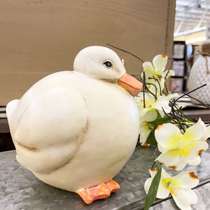 White ceramic duck figurine with a orange beak and feet, placed on a surface with yellow flowers.  Chubby White Resin Duck Figurine available at Quilted Cabin Home Decor.