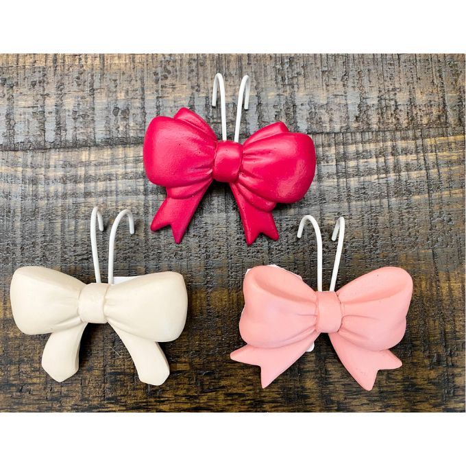 Three bow-shaped pot hangers in pink, white, and pink on a wooden surface. Bow Pot Hangers - Three Colours available at Quilted Cabin Home Decor.