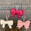 Three bow-shaped pot hangers in pink, white, and pink on a textured wooden surface. Bow Pot Hangers - Three Colours available at Quilted Cabin Home Decor.