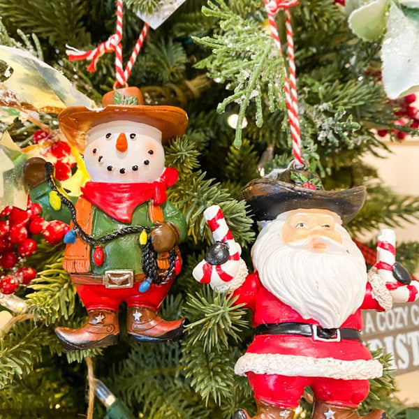 Two Christmas ornaments, a snowman and a Santa Claus figure, hanging on a decorated tree. Western Ornaments - Two Styles available at Quilted Cabin Home Decor.