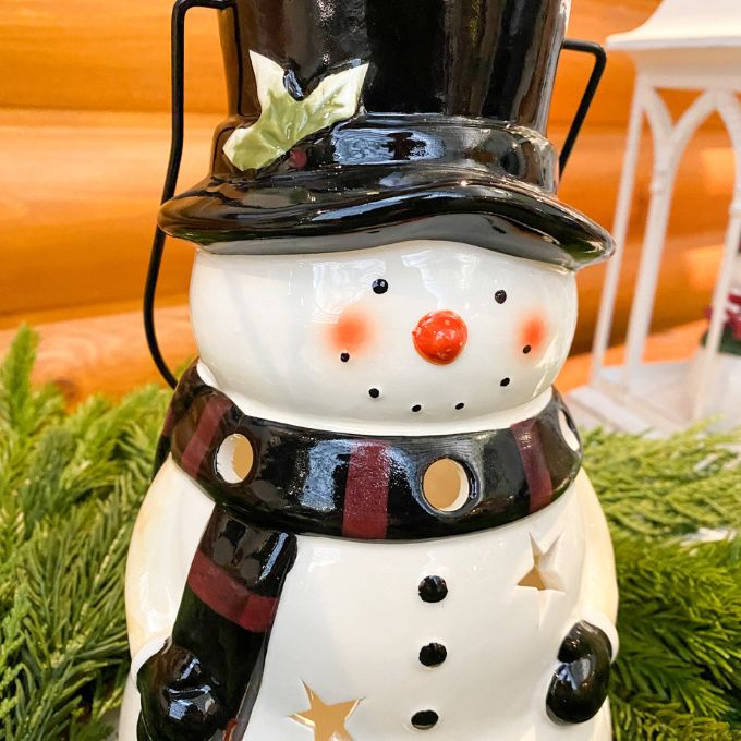 Decorative snowman lantern with a black top hat and scarf, surrounded by greenery. Snowman Lantern available at Quilted Cabin Home Decor.