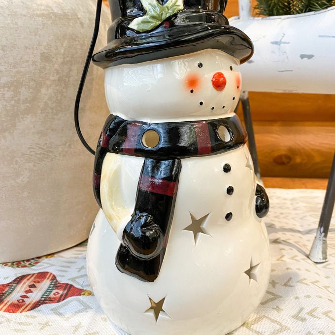 Decorative snowman with a black top hat and scarf on a patterned surface. Snowman Lantern available at Quilted Cabin Home Decor.