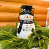 Snowman lantern with top hat and scarf on greenery against a wooden background. Snowman Lantern available at Quilted Cabin Home Decor.