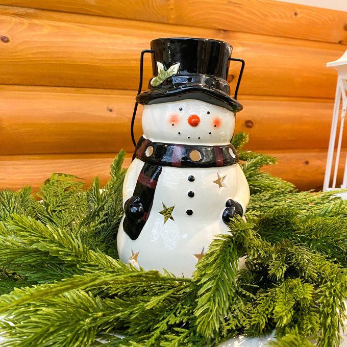 Snowman lantern with top hat and scarf on greenery against a wooden background. Snowman Lantern available at Quilted Cabin Home Decor.
