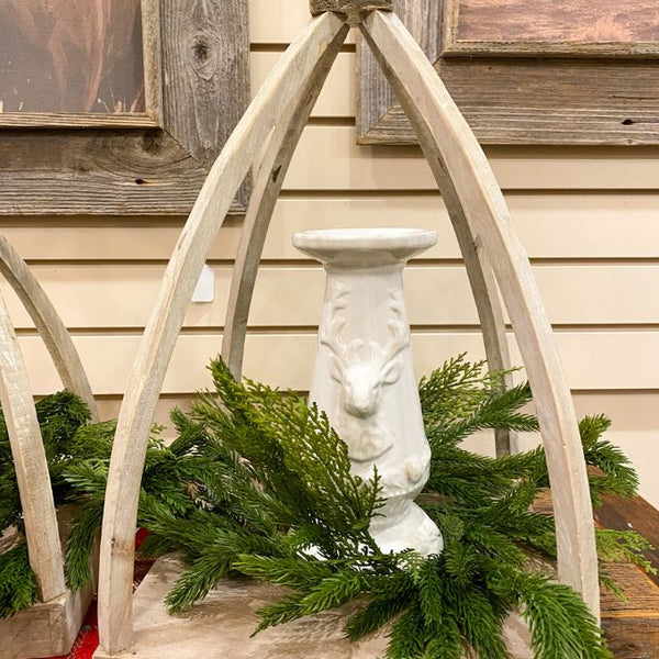 White ceramic deer embossed pillar candle holder  and greenery against a wooden wall. Christmas Deer Pillar Candle Holder available at Quilted Cabin Home Decor.