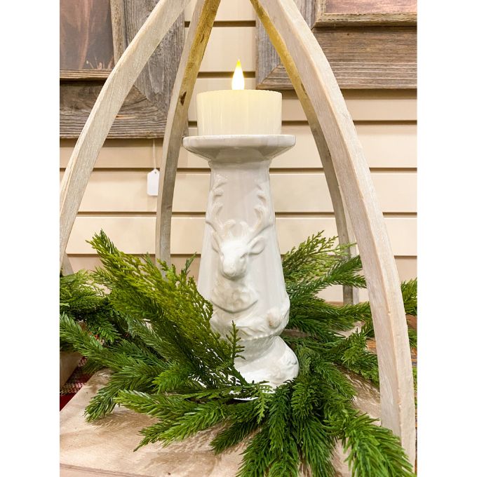 Decorative candle holder with a deer design surrounded by greenery against a wooden background. Christmas Deer Pillar Candle Holder available at Quilted Cabin Home Decor.