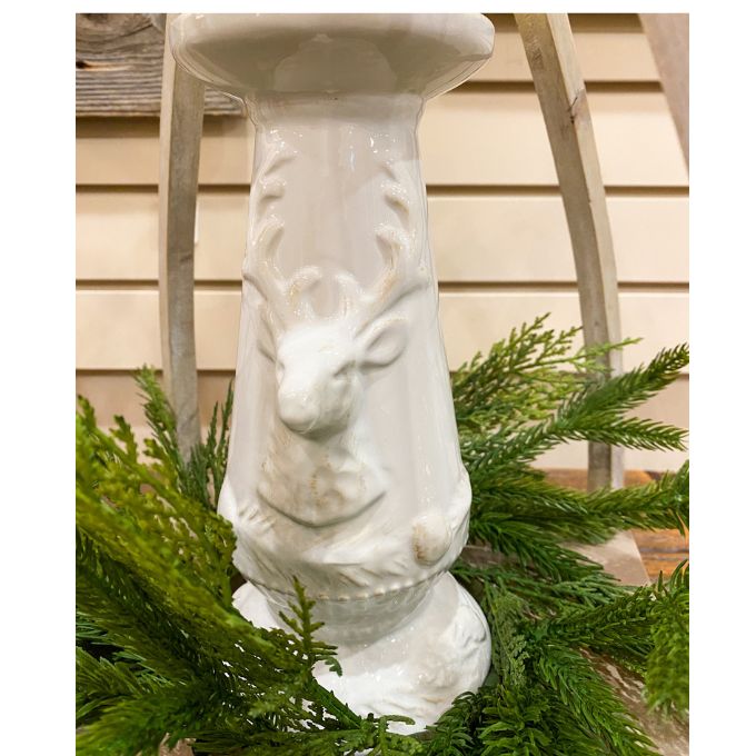 Decorative white pedestal with deer heads on a background of greenery. Christmas Deer Pillar Candle Holder available at Quilted Cabin Home Decor.