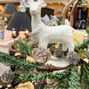 Decorative white deer figure on a table with greenery and pinecones in a store setting. Standing Christmas Deer available at Quilted Cabin Home Decor.
