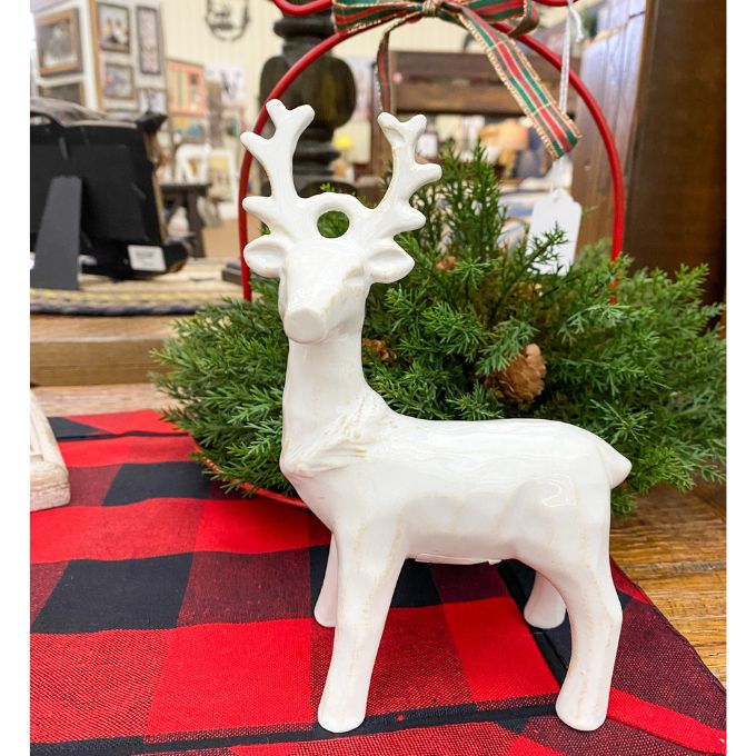 White ceramic deer figurine on a red and black checkered tablecloth with greenery in the background. Standing Christmas Deer available at Quilted Cabin Home Decor.