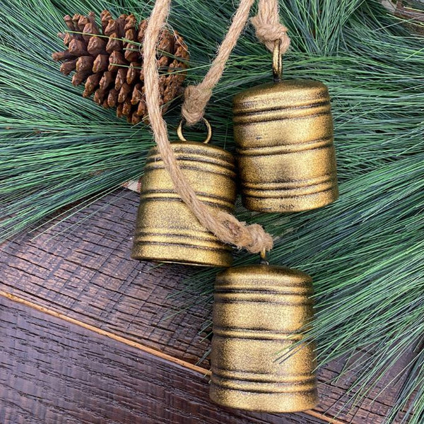 Three gold bells hanging on a jute string with pine cones and greenery on a wooden surface. Brushed Gold Bell Cluster available at Quilted Cabin Home Decor.