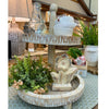 Decorative stone fountain with plants and vases in a store setting. Woodgrain Stoneware Flower Figurine available at Quilted Cabin Home Decor.