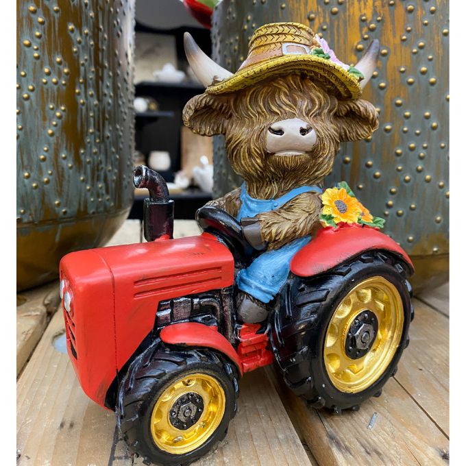 Figurine of a highland cow with a hat riding a red tractor on a wooden surface. Highland Cow Tractor Figurine available at Quilted Cabin Home Decor.