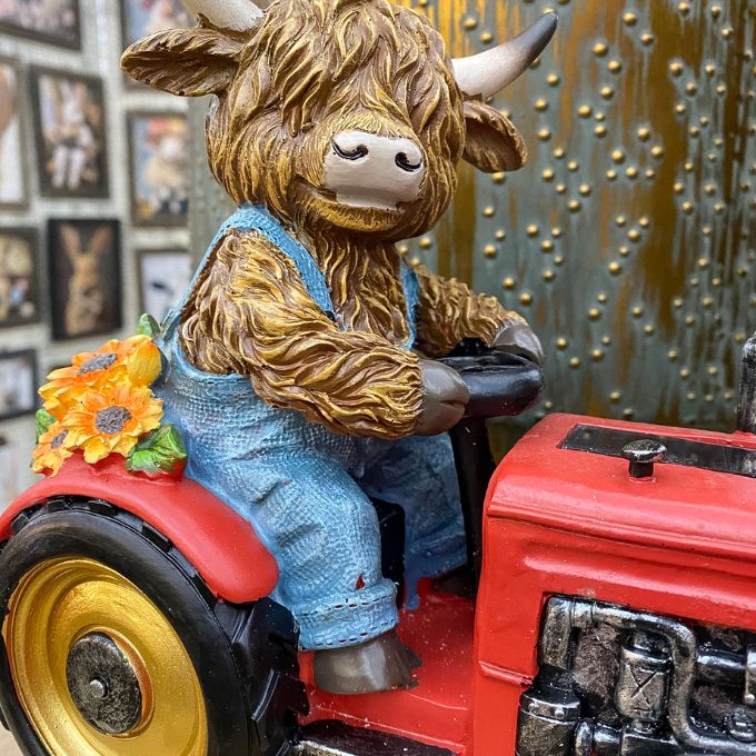 Figurine of a cow in overalls driving a red tractor with a sunflower decoration. Highland Cow Tractor Solar Figurine available at Quilted Cabin Home Decor.