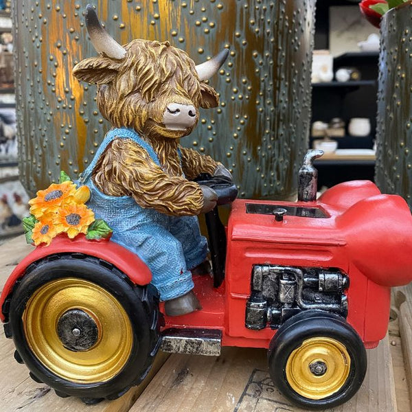 Figurine of a highland cow driving a red tractor with gold wheels on a wooden surface. Highland Cow Tractor Solar Figurine available at Quilted Cabin Home Decor.