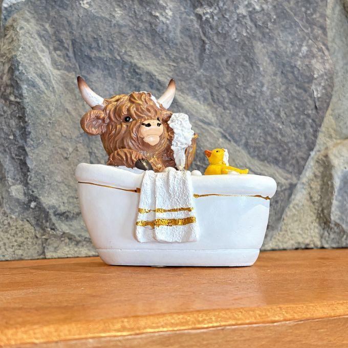 A decorative figurine of a brown Highland cow sitting in a white bathtub with gold trim, holding a white towel, with a small yellow rubber duck beside it. The scene is set on a wooden surface against a gray stone background.  Available at Rustic Ranch Furniture in Airdrie