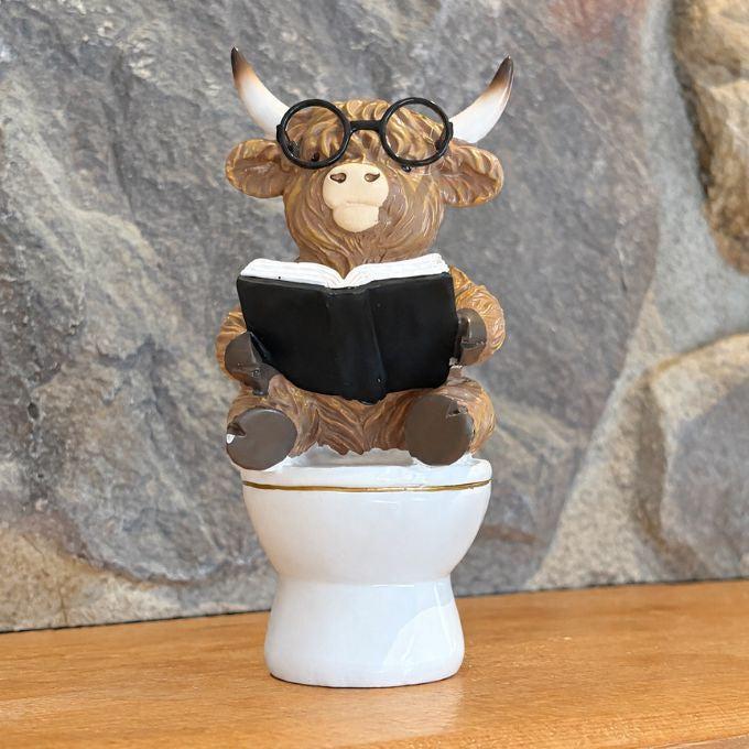 A decorative figurine of a shaggy brown bull wearing round black glasses and reading a black book while sitting on a white toilet, displayed on a wooden surface against a gray stone wall background.  Available at Quilted Cabin in Airdrie