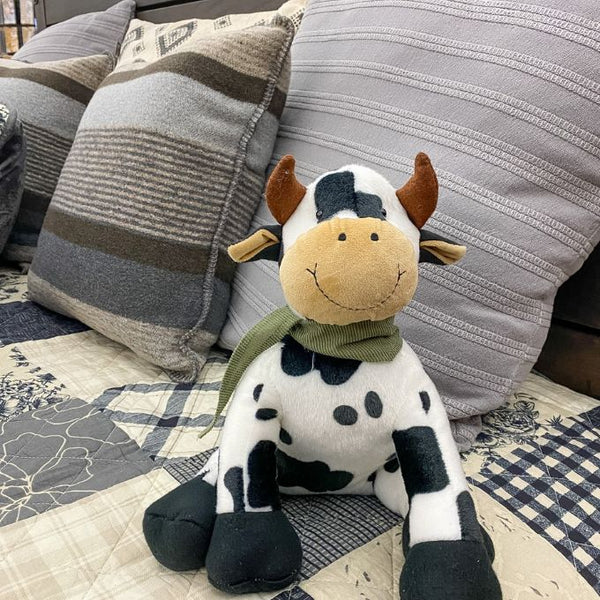 Plush cow toy on a patterned fabric background. Moo Cow Stuffie available at Quilted Cabin Home Decor.