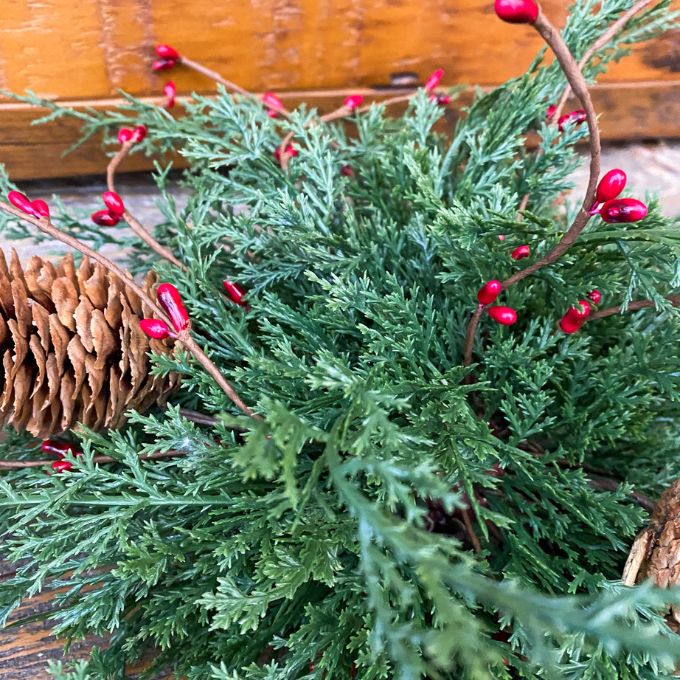 Green foliage with red berries and a pine cone on a wooden surface. Berry and Cedar Dripping Floral Orbs - Two Styles available at Quilted Cabin Home Decor.