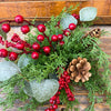 Christmas floral orb with red berries, greenery, and pine cones on a wooden background. Berry and Cedar Dripping Floral Orbs - Two Styles available at Quilted Cabin Home Decor.