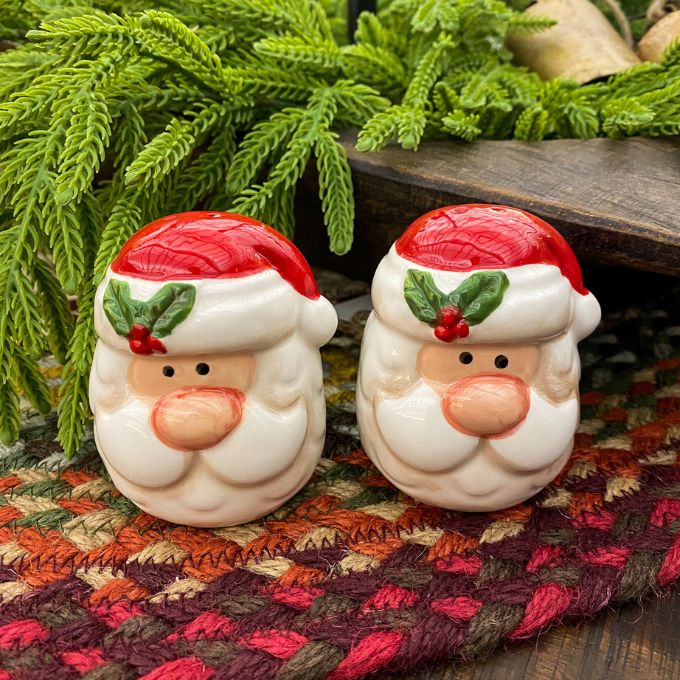 Two Santa Claus salt and pepper shakers on a textured surface with greenery in the background. Happy Santa Salt & Pepper Shakers available at Quilted Cabin Home Decor.