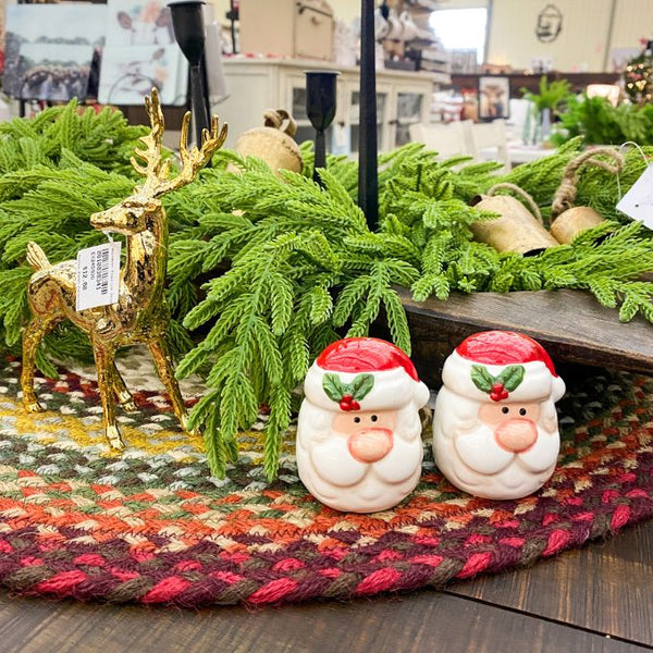 Santa Claus Salt and Pepper shakers with red hats and green holly leaves, a gold deer figurine, and greenery on a patterned rug. Happy Santa Salt & Pepper Shakers available at Quilted Cabin Home Decor.