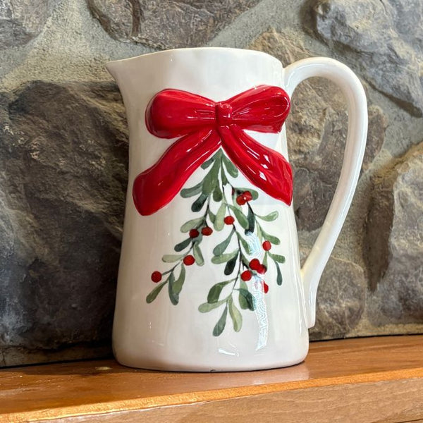 A white ceramic pitcher decorated with a large red bow and green holly branches with red berries, set against a rustic stone wall background. Available at Quilted Cabin in Airdrie