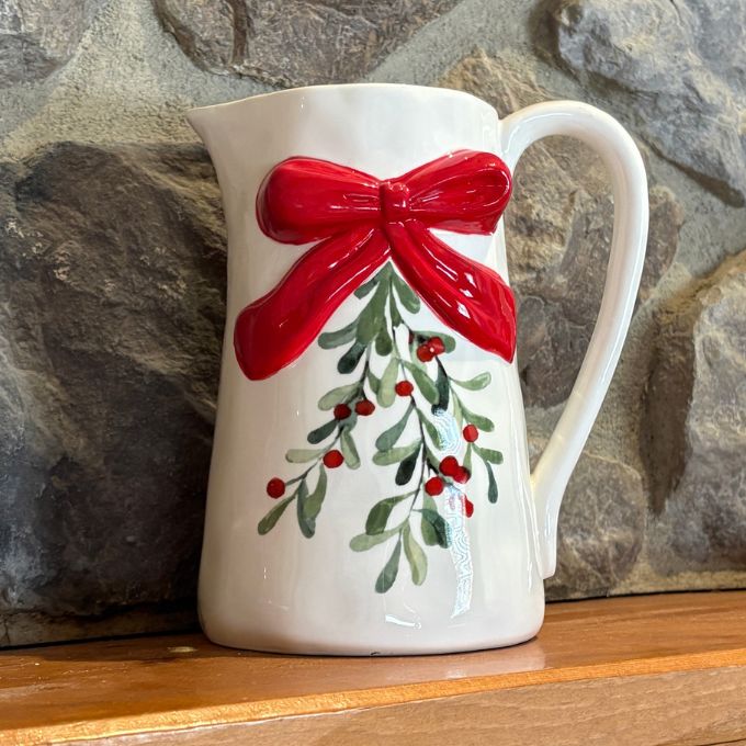 A white ceramic pitcher decorated with a large red bow and green holly branches with red berries, set against a rustic stone wall background. Available at Quilted Cabin in Airdrie