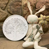 Decorative wall art with tree design next to a plush toy with bunny ears against a stone wall. Whitewashed Tree of Life Wall Art available at Quilted Cabin Home Decor.