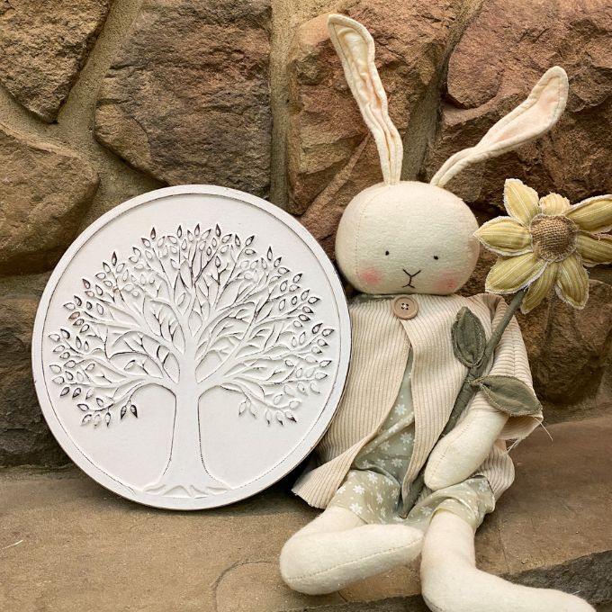 Decorative wall art with tree design next to a plush toy with bunny ears against a stone wall. Whitewashed Tree of Life Wall Art available at Quilted Cabin Home Decor.