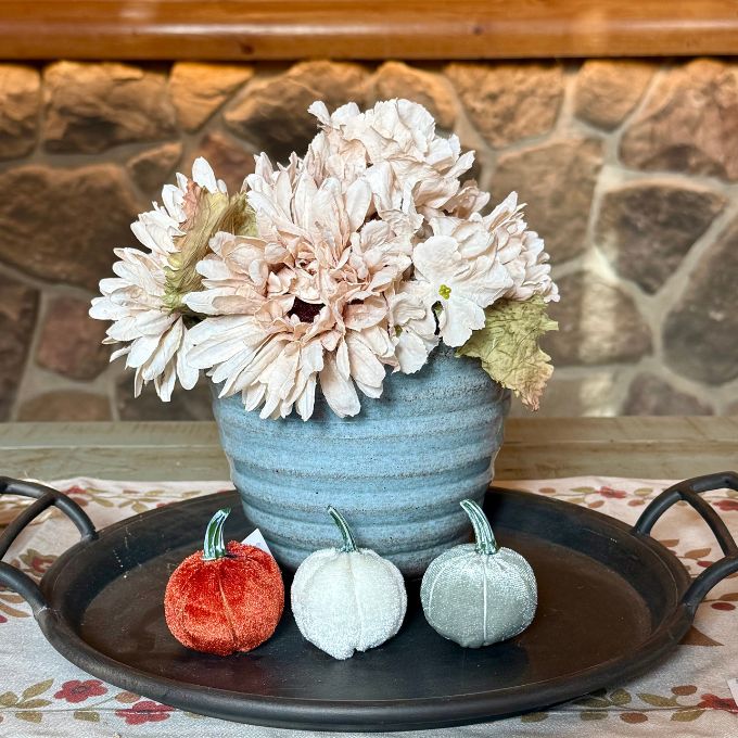 A decorative floral centerpiece in a ribbed light blue ceramic pot, styled on a black metal tray with three small velvet pumpkins in red, white, and sage green in front. The setup is on a floral tablecloth with a rustic stone wall background.  All items available at Quilted Cabin in Airdrie